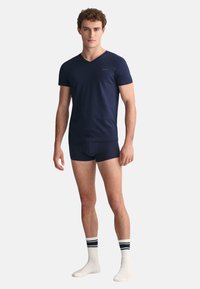 Navy Baumwoll-V-Ausschnitt-T-Shirt kombiniert mit passenden Boxershorts. Das Modell trägt außerdem weiße Sneakersocken mit navy und weißen Streifen. Einfaches Design, taillierte Form.