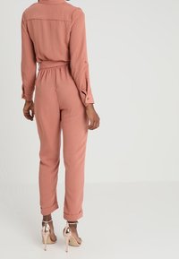 Persona vestida con un mono de manga larga color rosa empolvado, con cintura y puños elásticos, combinado con sandalias de tacón alto metálicas en dorado rosa, de espaldas.