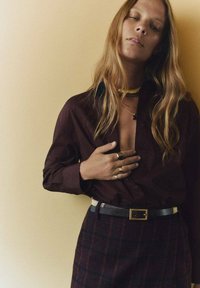 Dark brown button-up shirt, plaid skirt with dark red accents, black belt with gold buckle, layered gold necklaces, and rings on fingers.