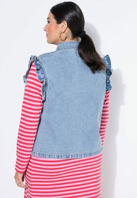 Lichtblauw denimvest met ruches op de schouders, gecombineerd met een langärmelig shirt met rood- en roze strepen. Toont een casual gelaagde stijl.