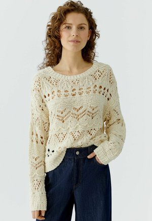 Vrouw met krullend haar, gekleed in een crèmekleurige, opengewerkte trui en donkere, hoogwater jeans, staand met één hand in haar zak tegen een witte achtergrond.