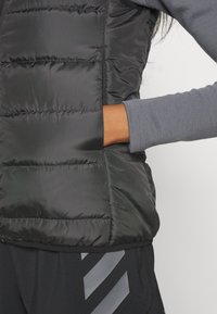 Juoda izoliacinė liemenė su siuvinėtu raštu, turinti vidutinio aukščio apykaklę, užtrauktukinius kišenes ir kontrastingą pilką ilgarankovę marškinėlį po ja.