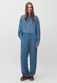 Denim kombinezon z dolgimi rokavi in gumbom, ki ima elastičen pas, široke hlače in svetlo modro barvo.