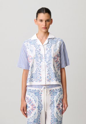 Donna che indossa una camicia e pantaloni bianchi abbinati con motivi geometrici intricati blu e rossi e maniche corte a righe blu, in piedi davanti a uno sfondo semplice.