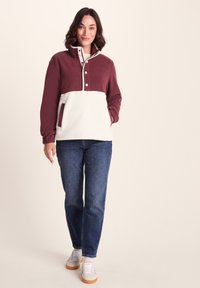 Woman wearing a maroon and white pullover fleece, blue jeans, and white sneakers, standing against a plain light background.
