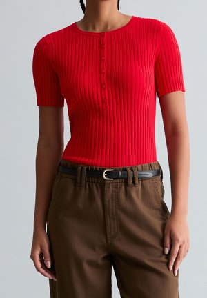 Kobieta ubrana w dopasowany czerwony prążkowany krótki rękaw z guzikami, włożony do brązowych spodni z wysokim stanem i czarnym paskiem.