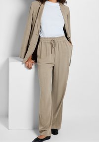 Béžový oblek s na míru šitým sakem a kalhotami s elastickým pasem, s hladkou texturou a volným střihem. Jednoduché světle modré tričko pod ním.