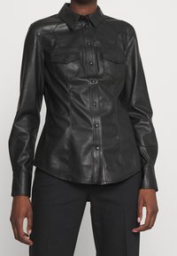 Camicia nera in pelle con colletto a punta, bottoni frontali e due tasche sul petto con flap. Texture liscia con design aderente.