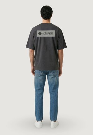 Uomo che indossa una maglietta Columbia Sportswear grigio scuro e jeans blu, in piedi di schiena con le braccia rilassate lungo i fianchi.