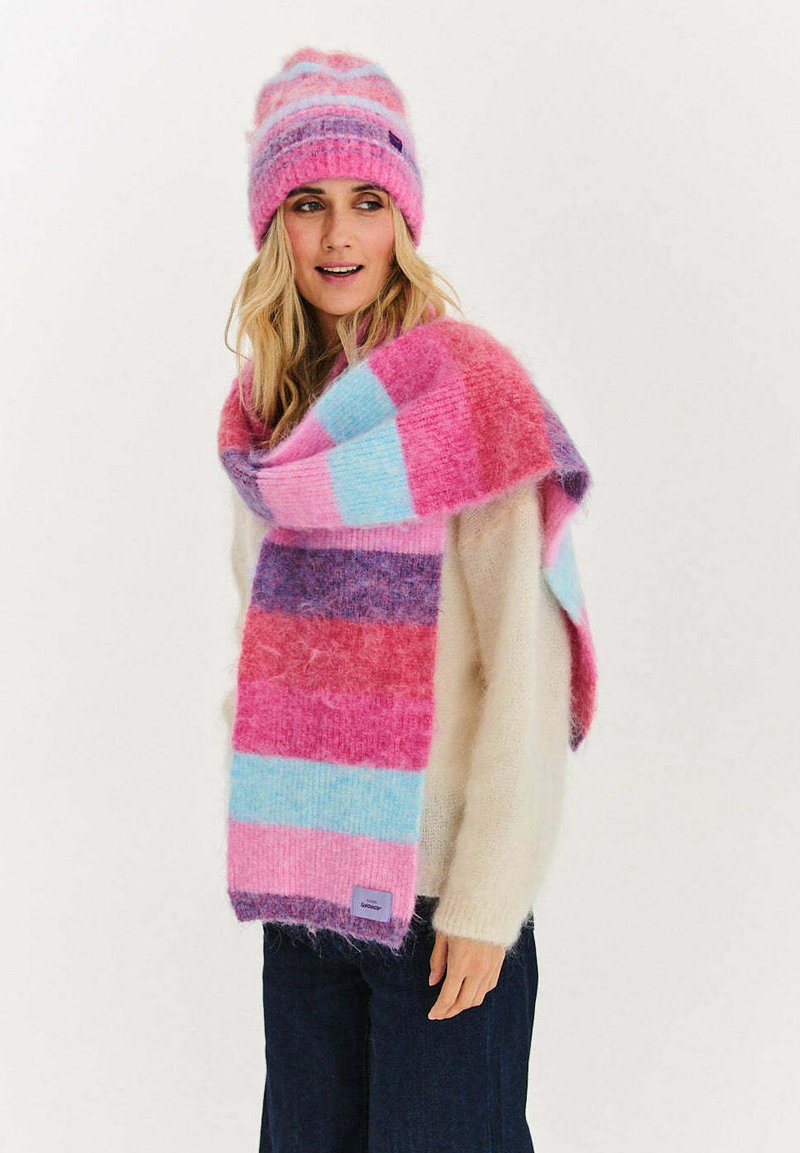 Różowo-niebieski szal w paski wykonany z miękkiej, puszystej tkaniny, ułożony na jasnym swetrze; dopasowana różowa czapka z wzorem w zygzak i metką marki.