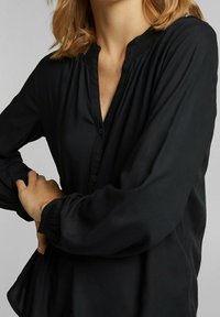 Sort button-up bluse med en afslappet silhuet, der har en opretstående krave, plissering omkring halsudskæringen og lange ærmer.
