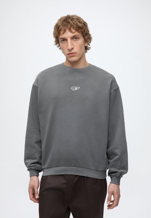 Mladić valovite kose u običnoj sivoj majici s okruglim izrezom i tamnim hlačama, stoji ispred bijele pozadine.