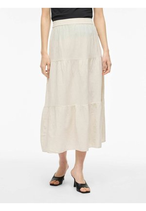 Vrouw draagt een lichtbeige midi-rok met gelaagde lagen en een elastische tailleband, gecombineerd met zwarte open-toe hakmuiltjes, tegen een witte achtergrond.