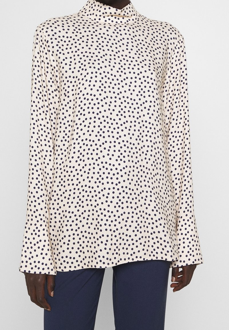 Blusa crema a maniche lunghe con pois navy indossata con pantaloni navy, mostrata dal collo ai fianchi su uno sfondo semplice.