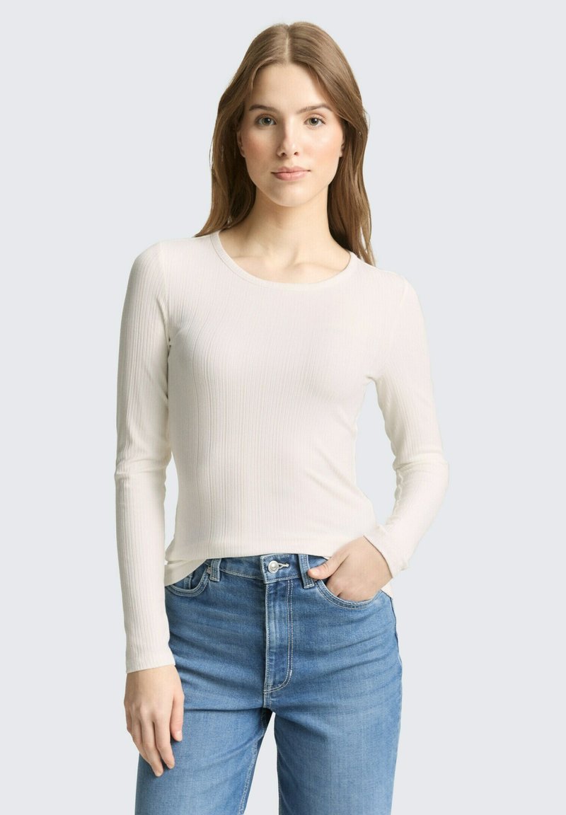 Jeune femme aux cheveux châtain clair lisses porte un haut blanc côtelé à manches longues ajusté et un jean bleu taille haute, main dans la poche.