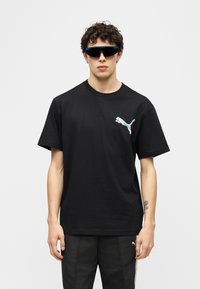 Νεαρός με σγουρά μαλλιά που φοράει μαύρο μπλουζάκι Puma, μαύρο παντελόνι και μεγάλα μαύρα γυαλιά ηλίου, με φόντο ένα απλό ανοιχτόχρωμο φόντο.