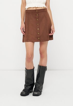 Frau trägt einen braunen Rock mit Knopfleiste vorne, ein weißes kurzes Oberteil und schwarze Stiefel mit Schnallen bis zur Mitte der Wade, steht vor weißem Hintergrund.