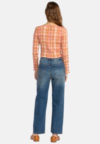 Femme avec de longs cheveux portant un haut à manches longues à carreaux orange, un jean bleu taille haute et des sabots marron, se tenant de dos sur un fond blanc.