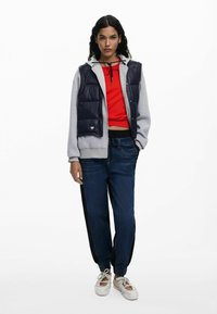 Gevoerde marineblauwe vest over een grijze hoodie, rode crop top en donkerblauwe joggingbroek met zwarte zijstrepen. Witte sandalen maken de outfit compleet.