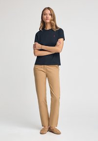 Kobieta stoi z założonymi rękoma, ubrana w czarną koszulę z krótkim rękawem, beżowe spodnie i beżowe buty wsuwane, na jednolitym tle.