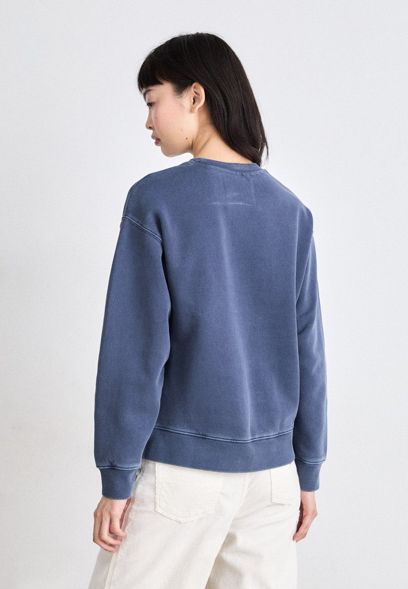 Bluza z okrągłym dekoltem w wyblakłym niebieskim kolorze bawełny. Posiada ściągacze przy mankietach i u dołu, luźny krój oraz subtelne detale wyszywane z tyłu.