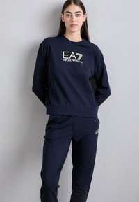 Mārciņā zilas sporta bikses un džemperis ar garām piedurknēm, kuru rotā zelta "EA7 EMPORIO ARMANI" logo un atbilstošas pieguļošas bikses. Mīksts audums un brīva piegriezuma forma.