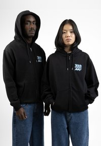 Zwarte rits-hoodies met voorzakken, voorzien van een blauw logo op de linkerkant van de borst. Deze zijn gecombineerd met blauwe spijkerbroeken.