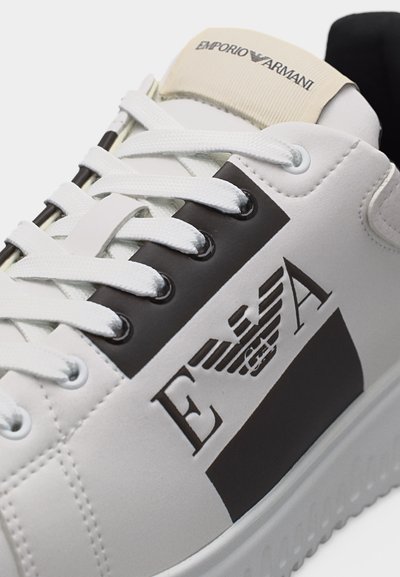 Λευκό αθλητικό παπούτσι με λευκά κορδόνια, μαύρο πάνελ με οπές, λογότυπο Emporio Armani στο πλάι και μπεζ ετικέτα στη γλώσσα με το όνομα της μάρκας.
