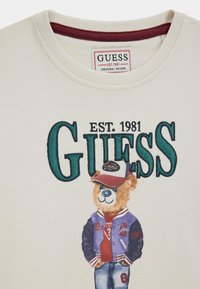 Bílý svetr s velkým zeleným textem "GUESS EST. 1981" a potiskem medvěda v baseballové čepici, varsity bundě a džínách pod textem.