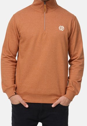 Mann trägt einen rostfarbenen Quarter-Zip-Pullover mit kleinem weißen Logo auf der linken Brust und schwarze Hose, Hände in den Taschen.