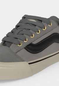 Baskets en toile grise avec des lacets gris épais, des œillets dorés, une bande latérale noire et une semelle en caoutchouc blanc cassé avec un liseré noir.