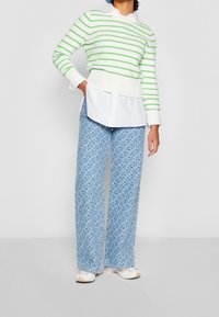 Zielono-biały sweter w paski nałożony na białą koszulę, zestawiony z niebieskimi spodniami w szerokich nogawkach w wzory oraz jasnymi trampkami.