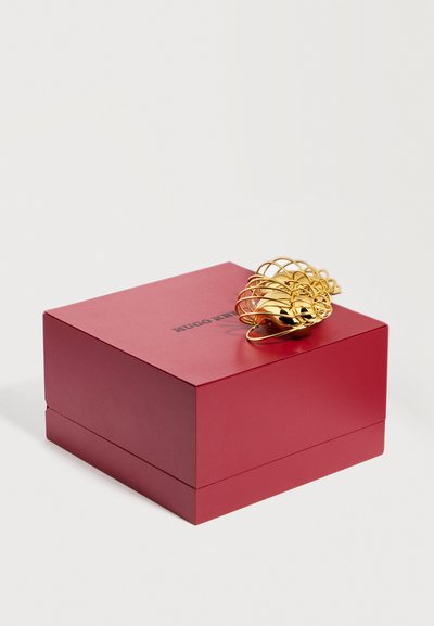 Κόκκινο κουτί Hugo Boss με μαύρο λογότυπο και ένα διακοσμητικό χρυσό αντικείμενο σε σχήμα κοχυλιού στην κορυφή, πάνω σε λευκό φόντο.