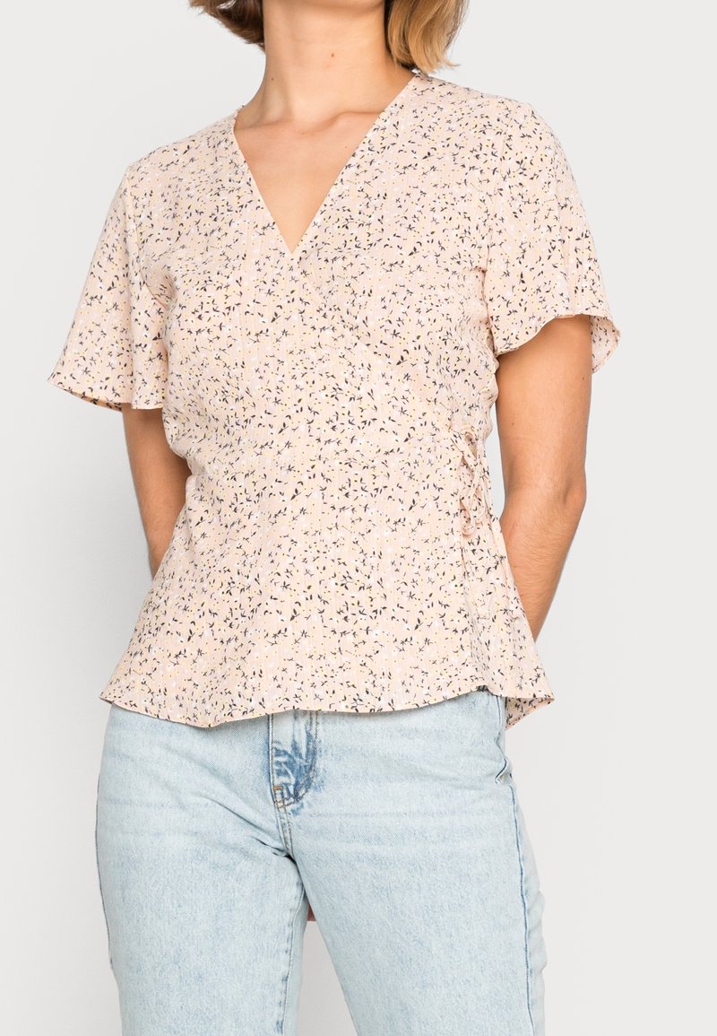 Femme portant un chemisier cache-cœur à manches courtes à fleurs rose pâle, associé à un jean taille haute bleu clair, debout devant un fond uni.