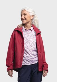 Giacca impermeabile rossa con cappuccio, dotata di zip frontale, indossata sopra una camicia leggera a pois rosa. I pantaloni cargo scuri completano l'outfit.