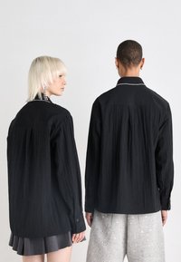 Camisas de manga longa negras texturizadas com punhos abotoados; uma delas tem um detalhe de colarinho com contas, ambas usadas com calças com padrão metálico.