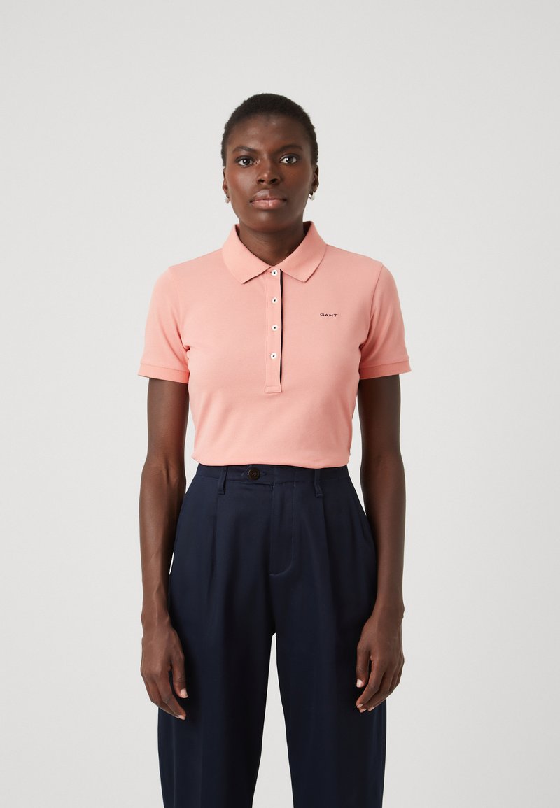 Polo shirt rosa chiaro con colletto, maniche corte e patta con cinque bottoni. Presenta un piccolo logo sul lato sinistro del petto. Indossato con pantaloni a vita alta blu navy.