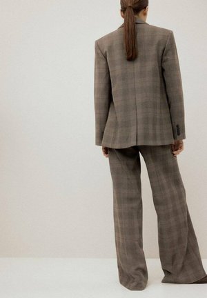 Personne aux longs cheveux bruns attachés en queue de cheval, portant un grand blazer à carreaux marron et un pantalon large assorti, debout face à un mur uni.