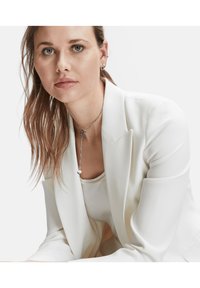 Weißer Blazer mit glatter Textur, getragen über einem hellen Oberteil; silberne Ketten Halskette mit rundem Anhänger, sichtbare Ohrringe und glattes Haar.
