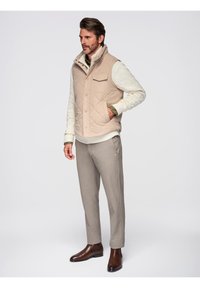 Gilet trapuntato beige sopra un maglione cremisi a trama; pantaloni grigi e stivali di pelle marroni. Presenta una tasca laterale sul gilet. Palette di colori neutri.
