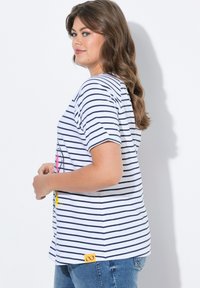 Pruhované tričko v bílé a námořnické barvě, s krátkými rukávy a volným střihem. Žlutá etiketa na spodním levém rohu. Nošeno s džínami.