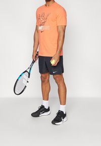 Mies, joka seisoo urheiluvaatteissa, kädessään tennismaila ja tennispallo. Hänellä on päällään oranssi Nike-t-paita, mustat Nike-shortsit ja mustat Nike-kengät.