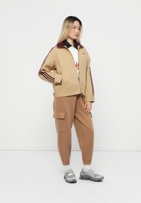 Giacca beige con zip e strisce bordeaux, abbinata a pantaloni della tuta marroni con tasche cargo. Indossata con scarpe da ginnastica grigie.