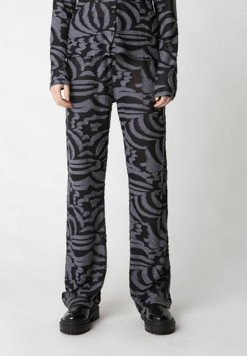 Schwarz gemustertes Set mit langärmeliger Bluse und weit geschnittenen Hosen. Strukturierter grauer Blumenprint auf dem Stoff. Stilisiert mit klobigen schwarzen Schuhen.