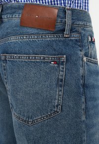 Tamsiai mėlynos džinsinės kelnės su ruduo odiniu etiketė "TOMMY HILFIGER." Ant užpakalinio kišenės yra mažas raudonas, baltas ir mėlynas logotipas.