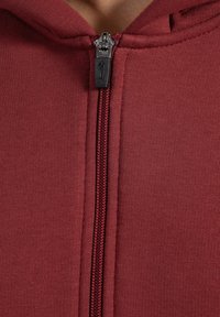 Sudadera burdeos con una textura de tejido suave, que presenta una cremallera de longitud completa en un color metálico contrastante y capucha.