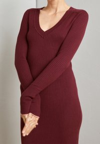 Burgundy ribbad klänning i sweaterstil med V-ringning och långärmar med tumhål, tillverkad av strukturerad stickad tyg, figurnära silhuett.