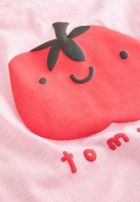 Grafika červeného rajčete na růžové tkanině, zobrazující usmívající se tvář s černými detaily. Pod ní je nápis „tom“ napsaný červenými zaoblenými písmeny.