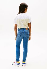 Camiseta blanca con rayas amarillas, jeans ajustados de mezclilla azules y zapatillas blancas con acentos azules y amarillos. Logo de Tommy Hilfiger visible.