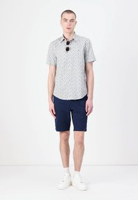 Kortermet hvit skjorte med grønt blomstermønster, sammen med marineblå shorts. Modellen har på seg hvite sneakers og solbriller, står foran en nøytral bakgrunn.